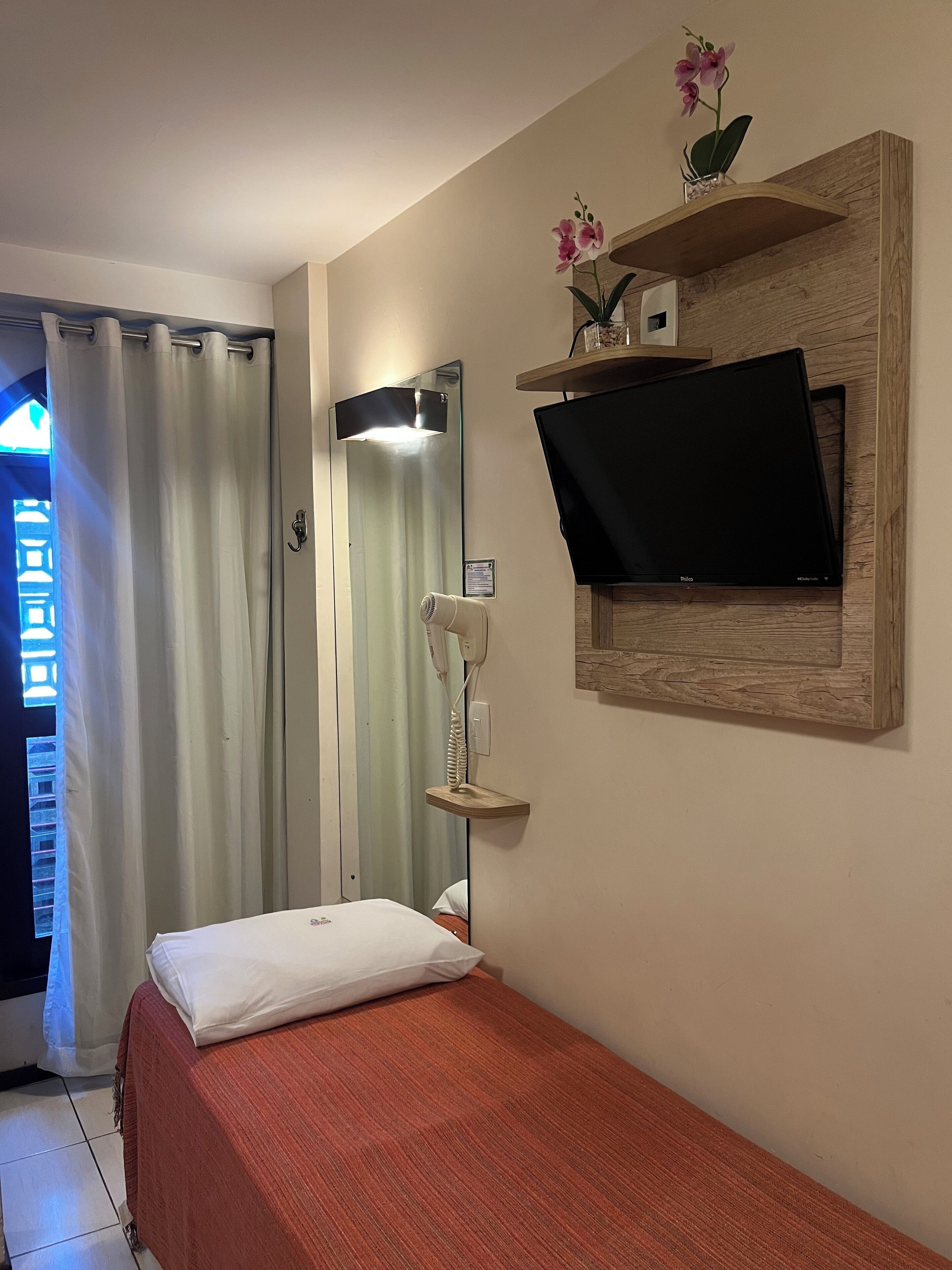 Habitación de hotel con cama, televisor montado en un estante de madera, secador de pelo sobre un soporte, espejo y ventana con cortinas.