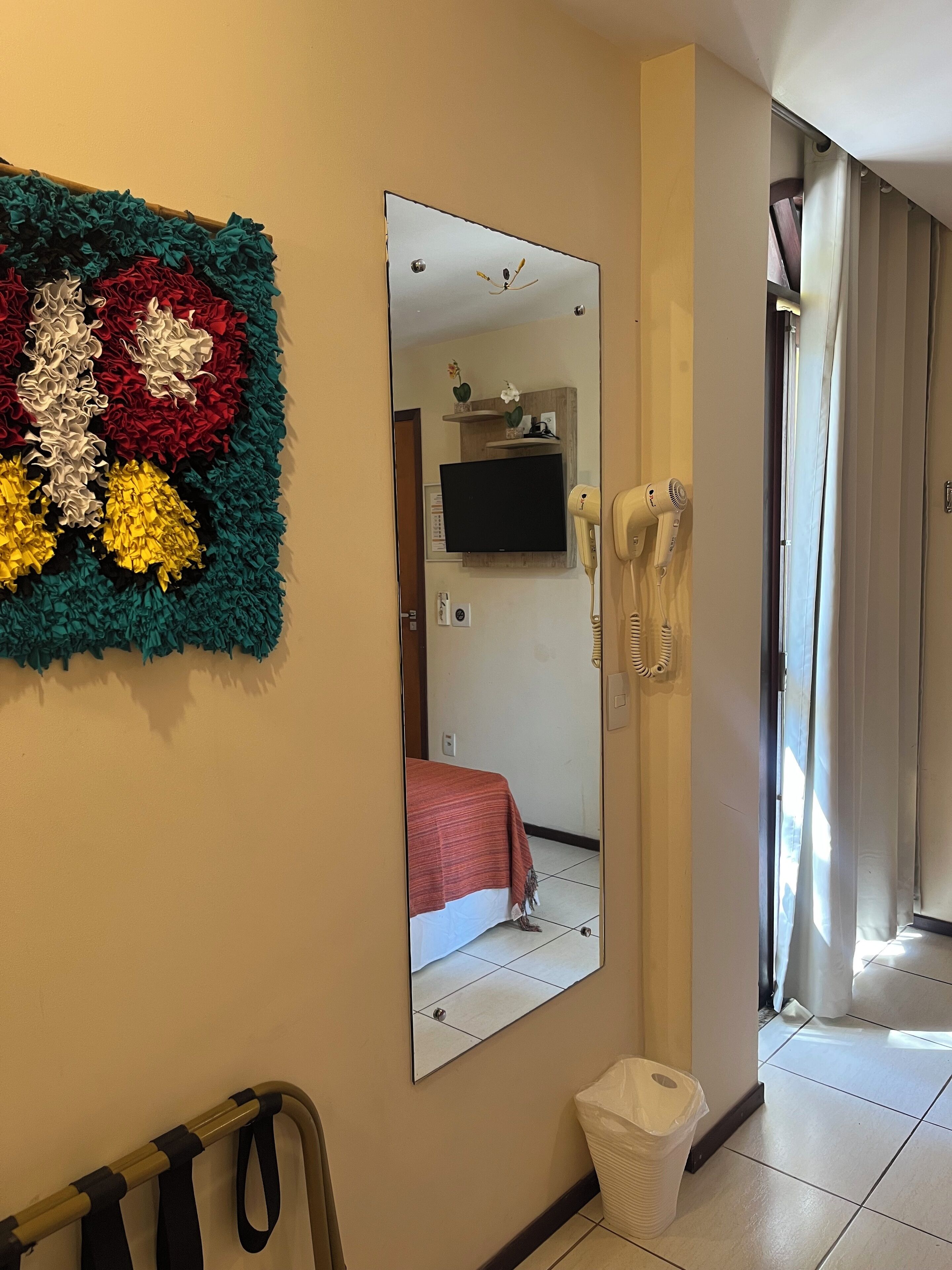 Habitación de hotel con un espejo grande, un televisor de pared, un secador de pelo y un cesto de basura.