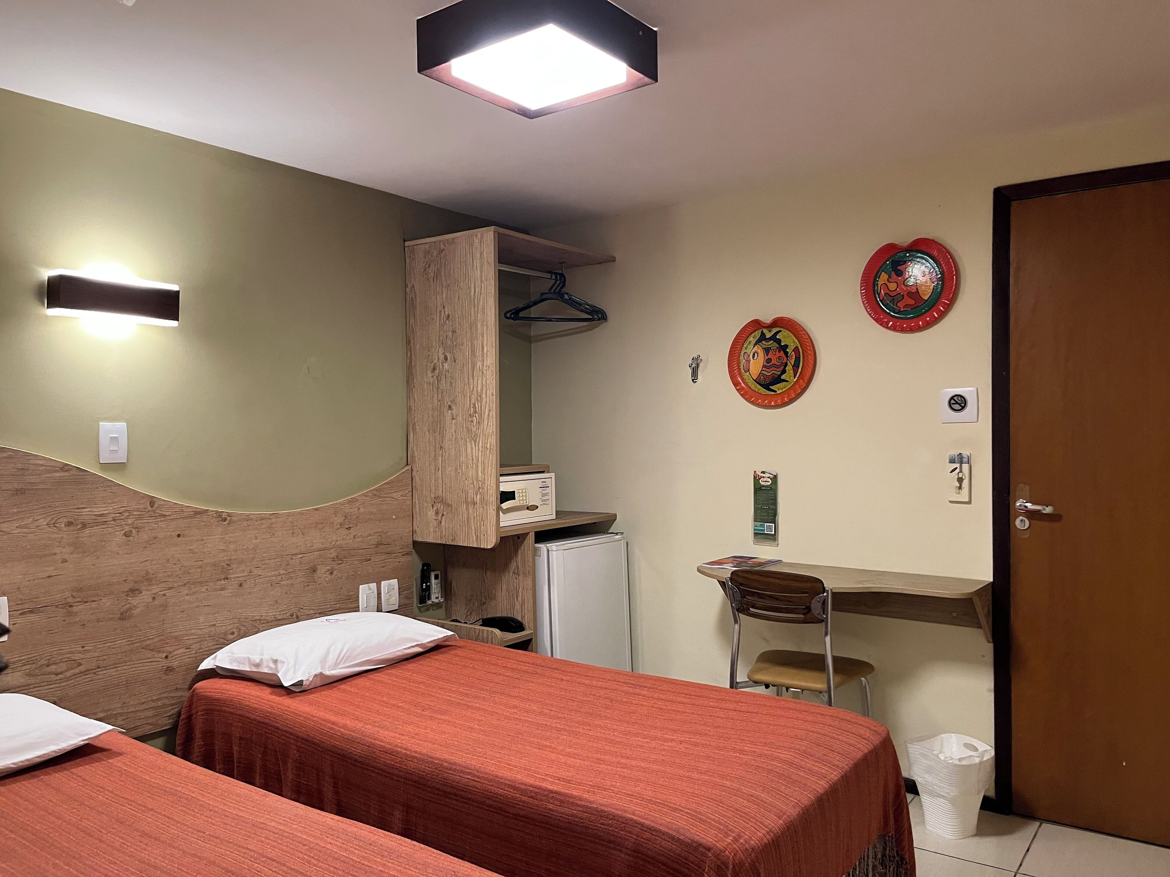 A hotel room with two beds, a desk, a chair, a small refrigerator, a trash bin, and a door.