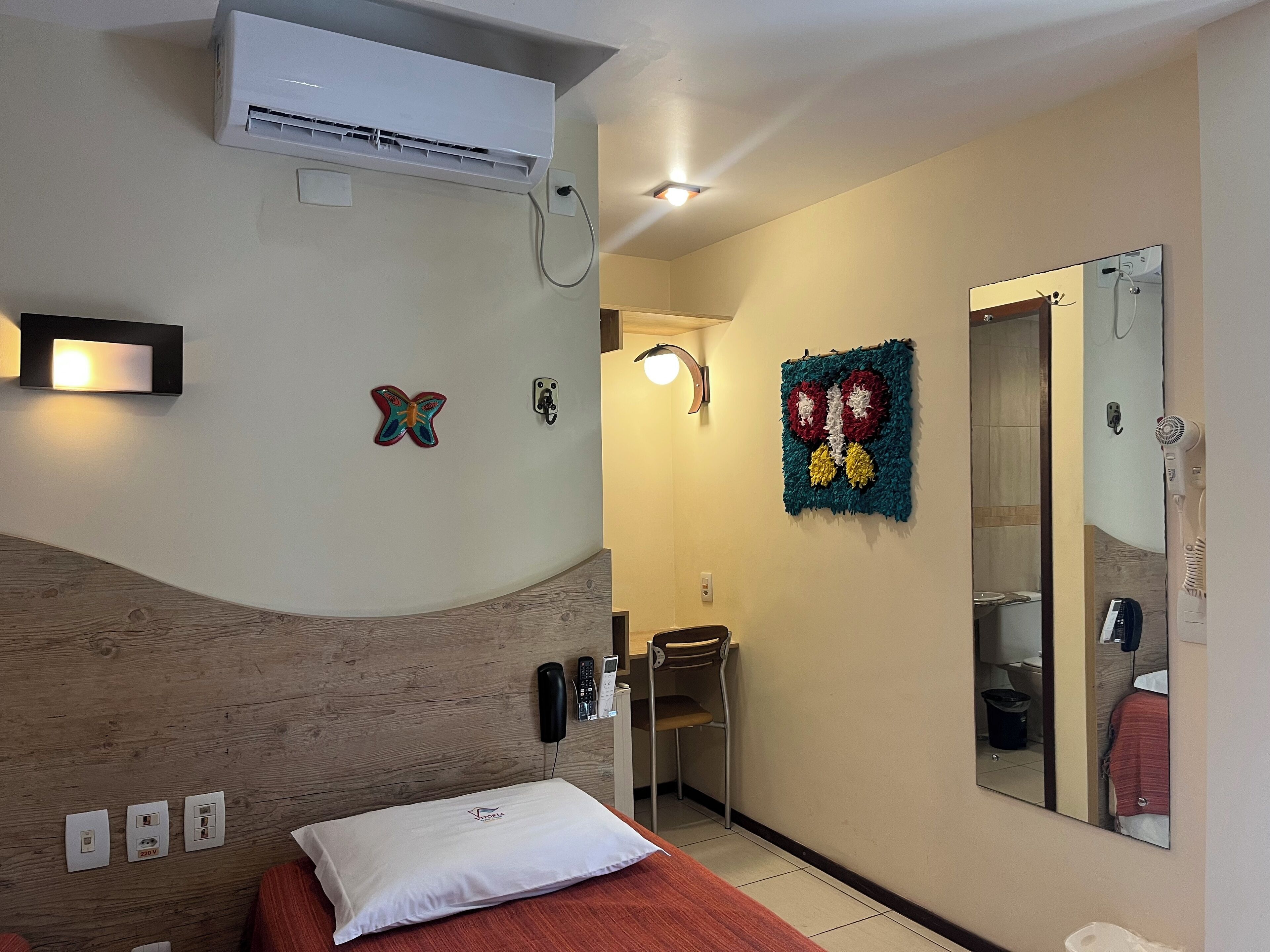 A hotel room with a bed, a bedside table, a mirror, a wall-mounted air conditioner, a wall-mounted phone, and a decorative butterfly on the wall.