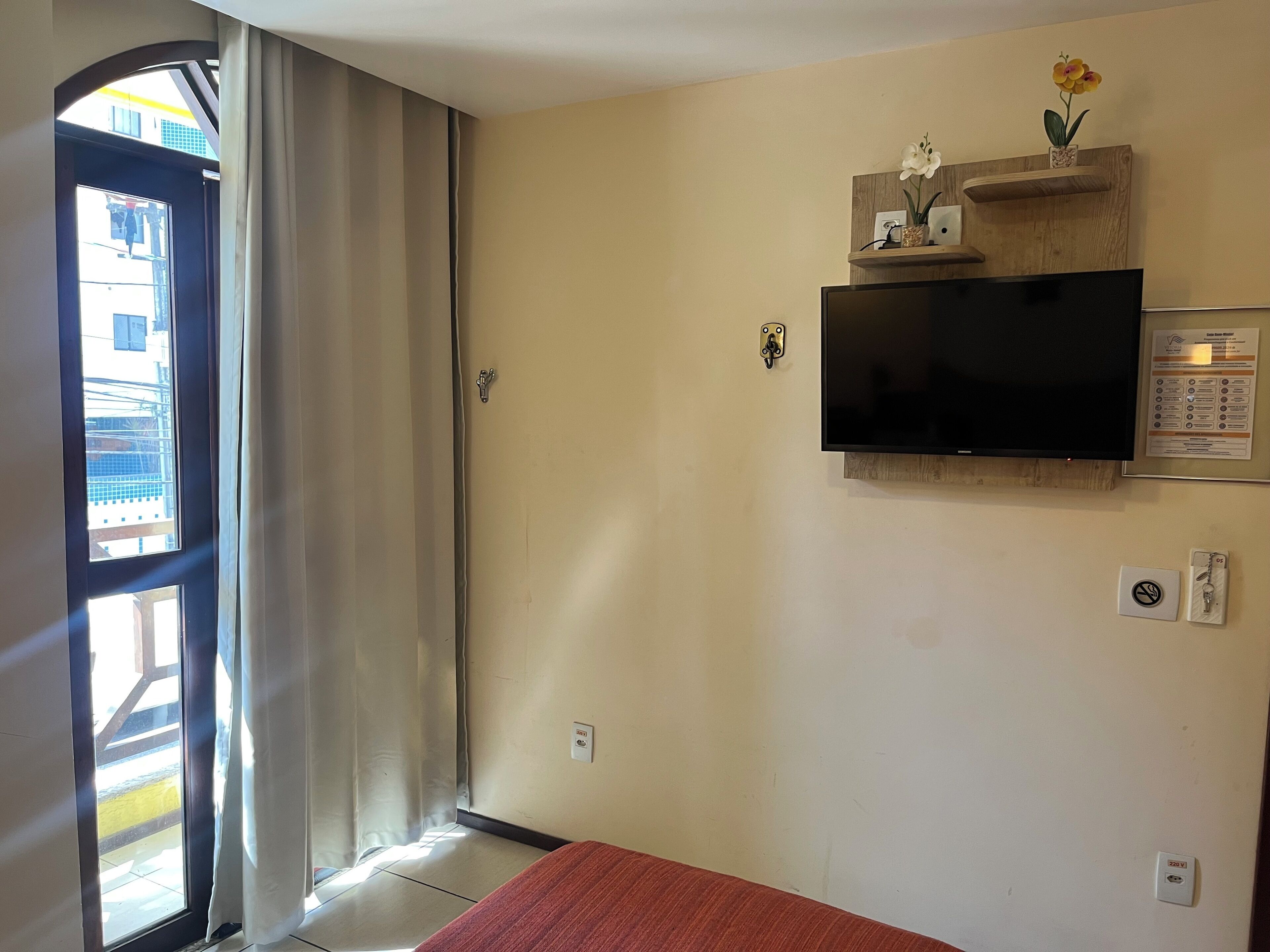 Habitación de hotel con un ventanal grande, un televisor de pantalla plana montado en la pared y una pequeña estantería con adornos.