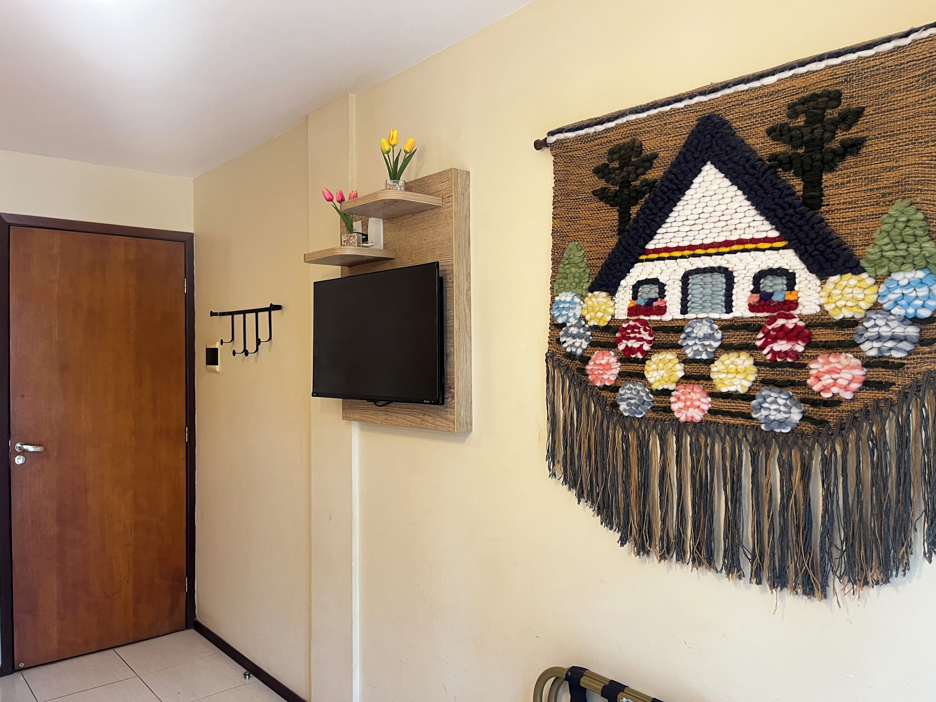 Una habitación con puerta de madera, un televisor de pantalla plana montado en la pared, un estante con flores y un tapiz tejido y colorido.