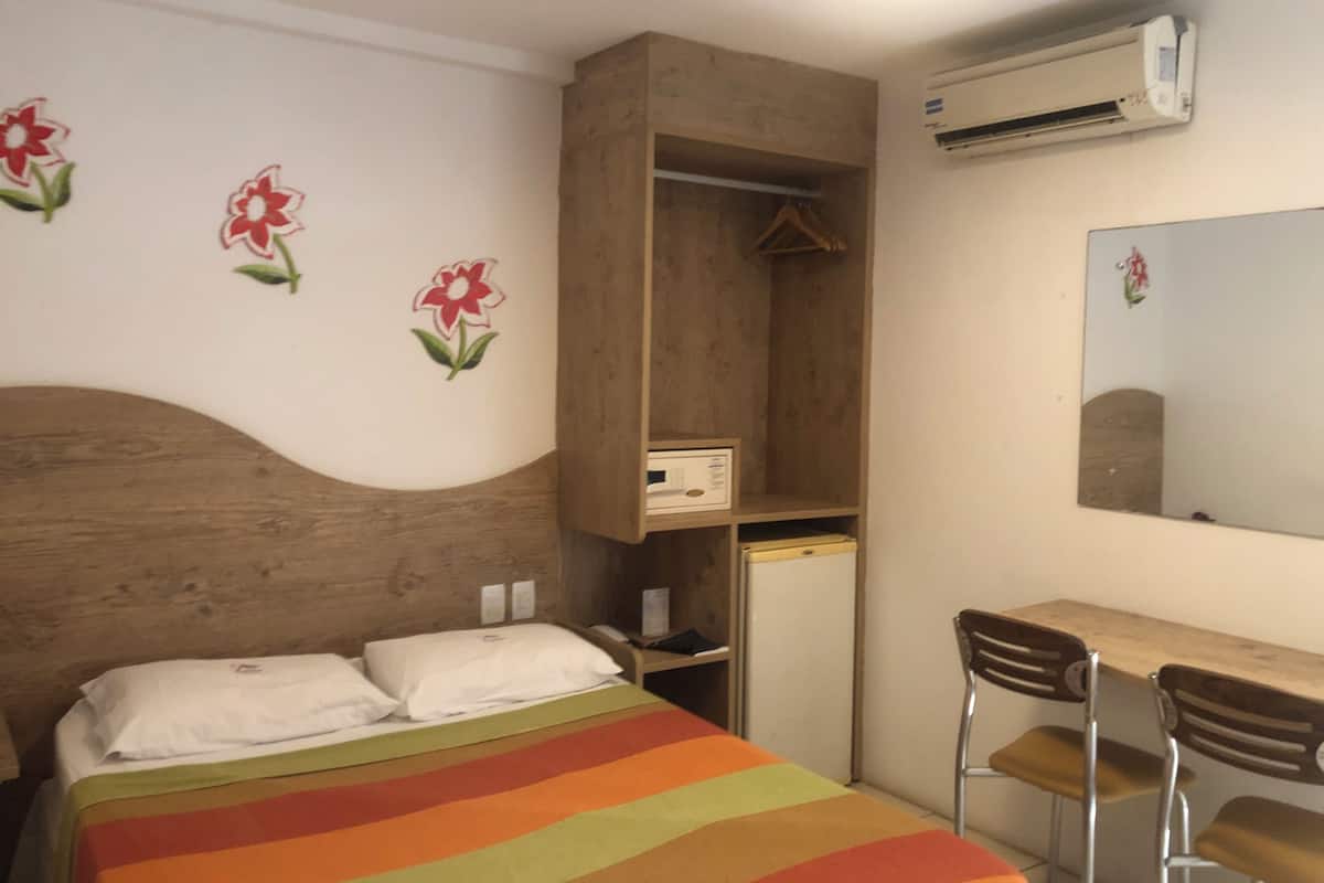 A bedroom with a bed, a desk, and a chair. There is a wardrobe, an air conditioning unit, and a mirror on the wall.