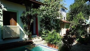 A pool area with a small staircase leading to a door, surrounded by lush greenery and a building with a tiled roof. - Irradiante (Olinda)