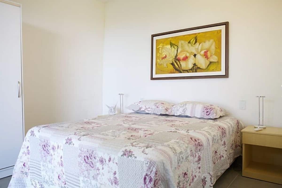 Un dormitori amb un llençol floral, una taula de nit amb una làmpada i un quadre amb flors al mur.