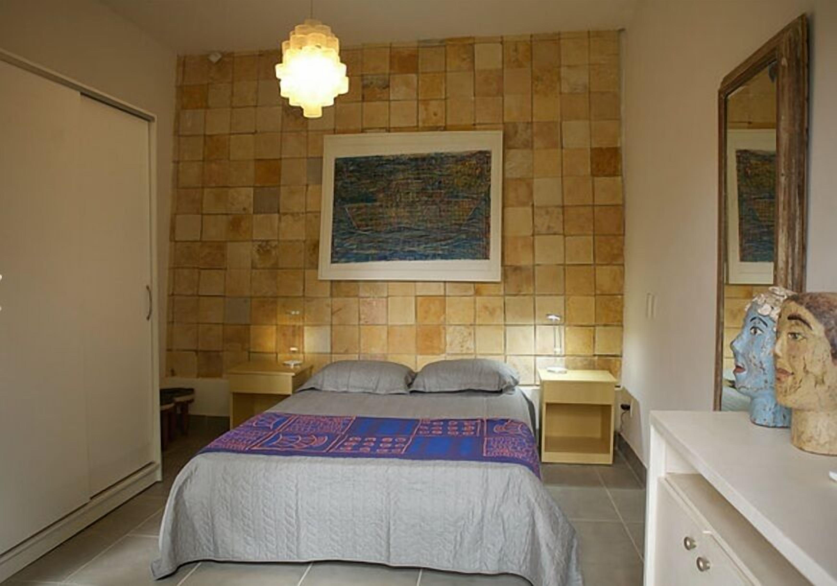 Un dormitorio con cama, mesitas de noche, un cuadro colgado en la pared y un aparador con objetos decorativos.