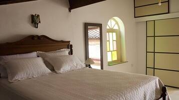 A bedroom with a wooden bed, white bedding, a nightstand, a wall-mounted lamp, and a window with a view.