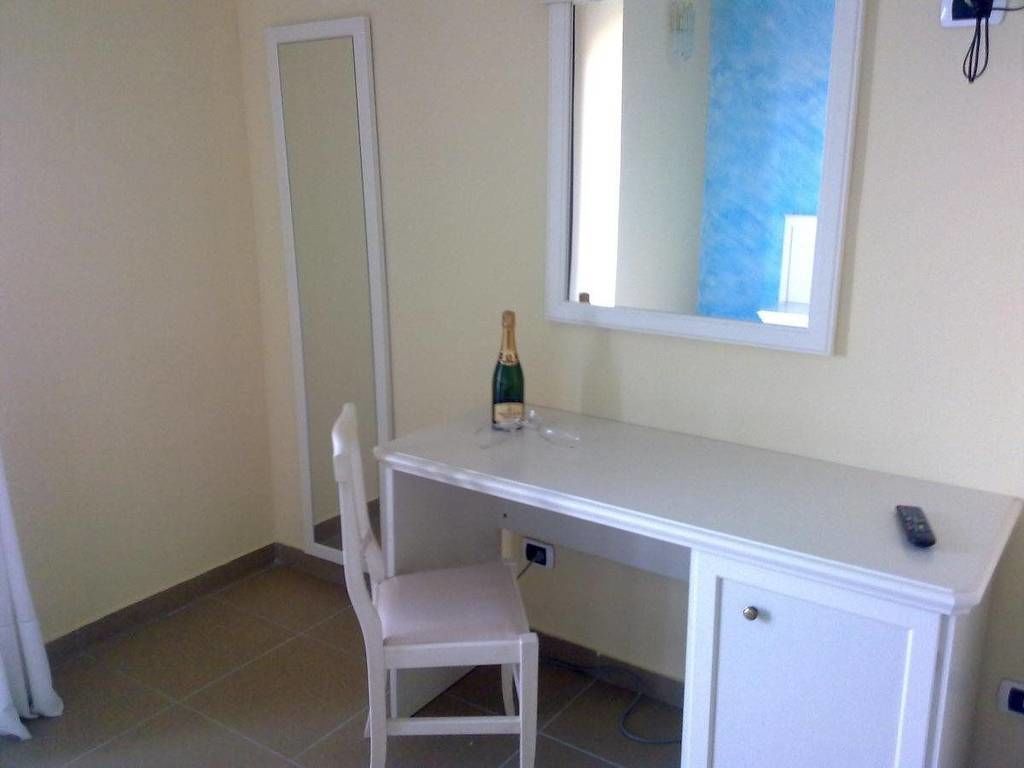 A small, clean room with a white desk, a chair, a bottle of champagne, and a mirror on the wall.