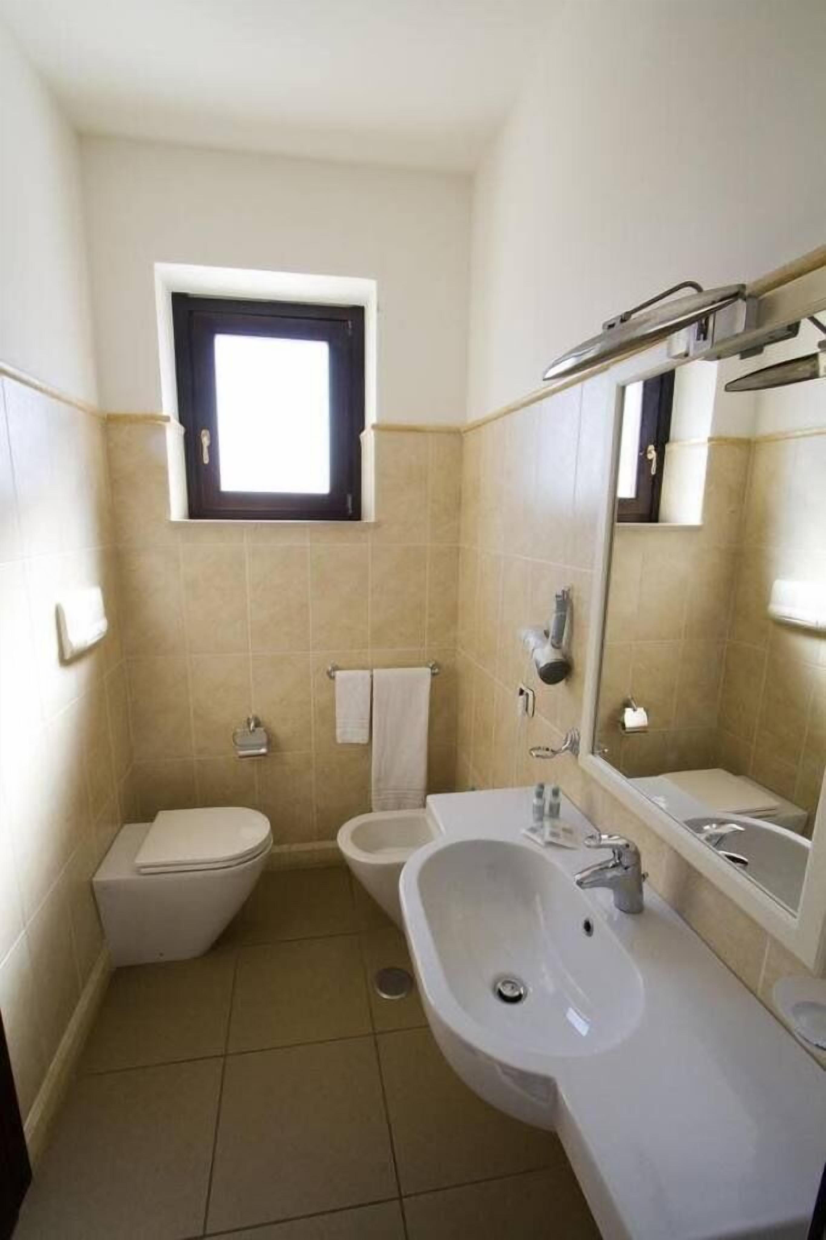 A small, clean bathroom with a toilet, bidet, and sink. There is a window above the toilet and a mirror above the sink.