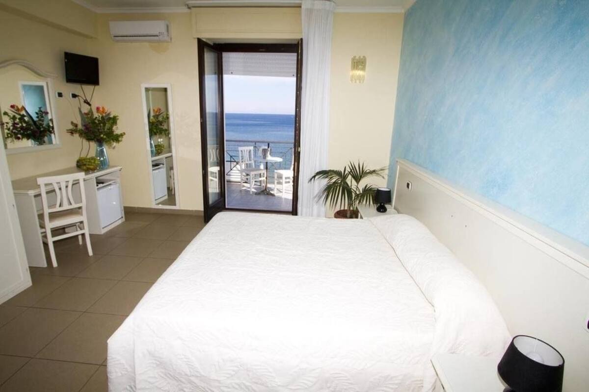 A hotel room with a large bed, a desk, a chair, a TV, a potted plant, and a view of the sea through a balcony door.
