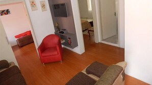 A living room with a red chair, a sofa, a TV on a wall unit, and a doorway leading to another room. - Apartamento dos Altos Centro de Gramado (Gramado)