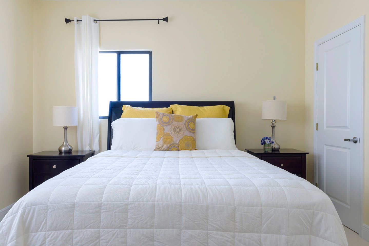 Una cama bien hecha con edredón blanco y dos almohadas, una ventana con vistas y una puerta a la derecha.