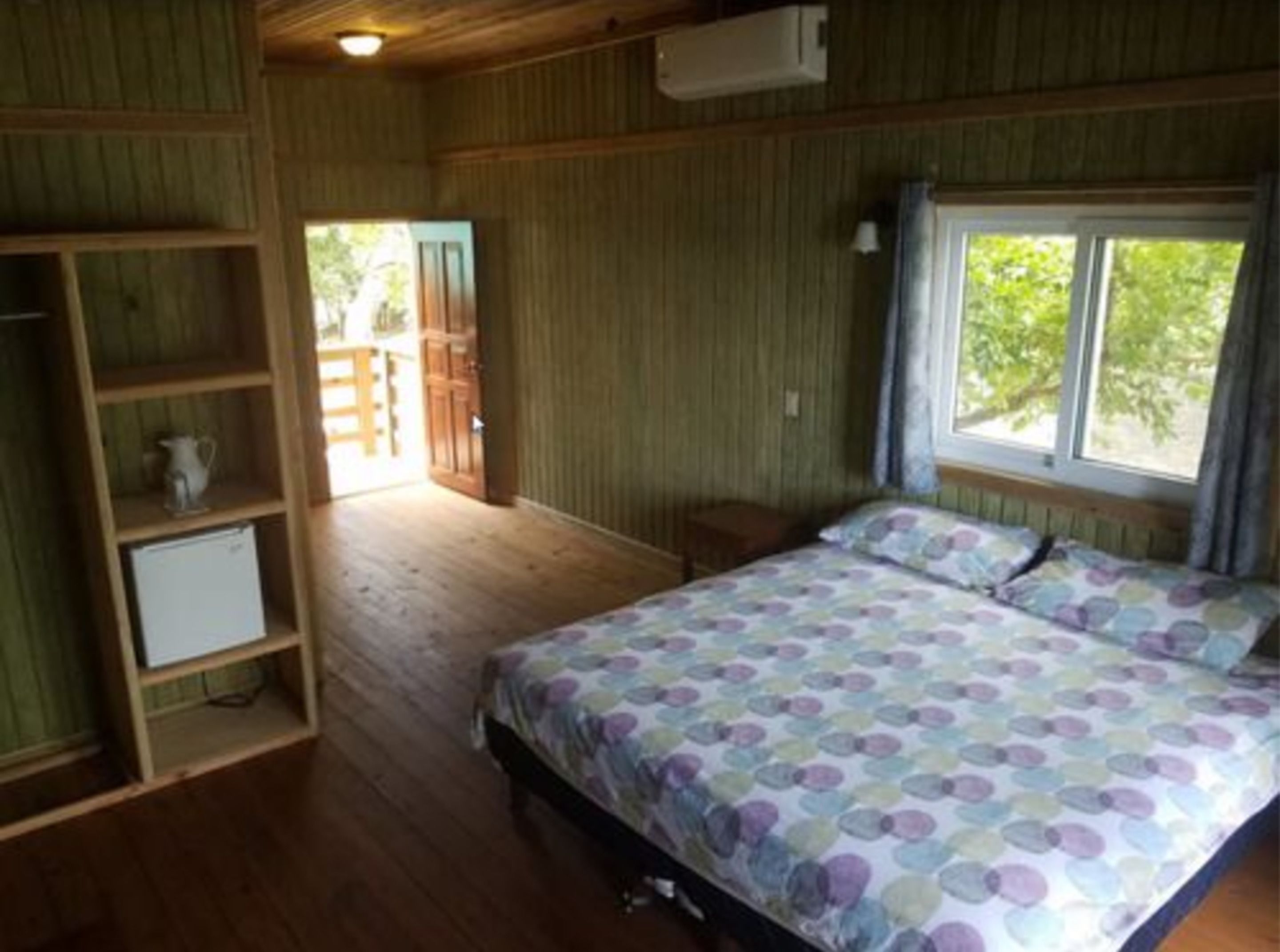 A bedroom with a bed, wooden shelves, a small table, and a window with a view of greenery.
