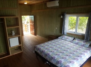 A bedroom with a bed, wooden shelves, a small table, and a window with a view of greenery. - Half Moon Resort (Roatan)