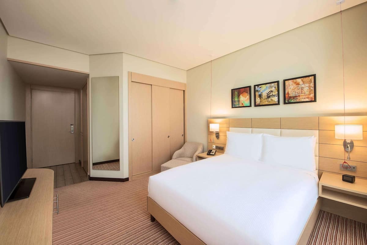 A hotel room with a bed, bedside tables, a chair, a desk, a wardrobe, and artwork on the wall.