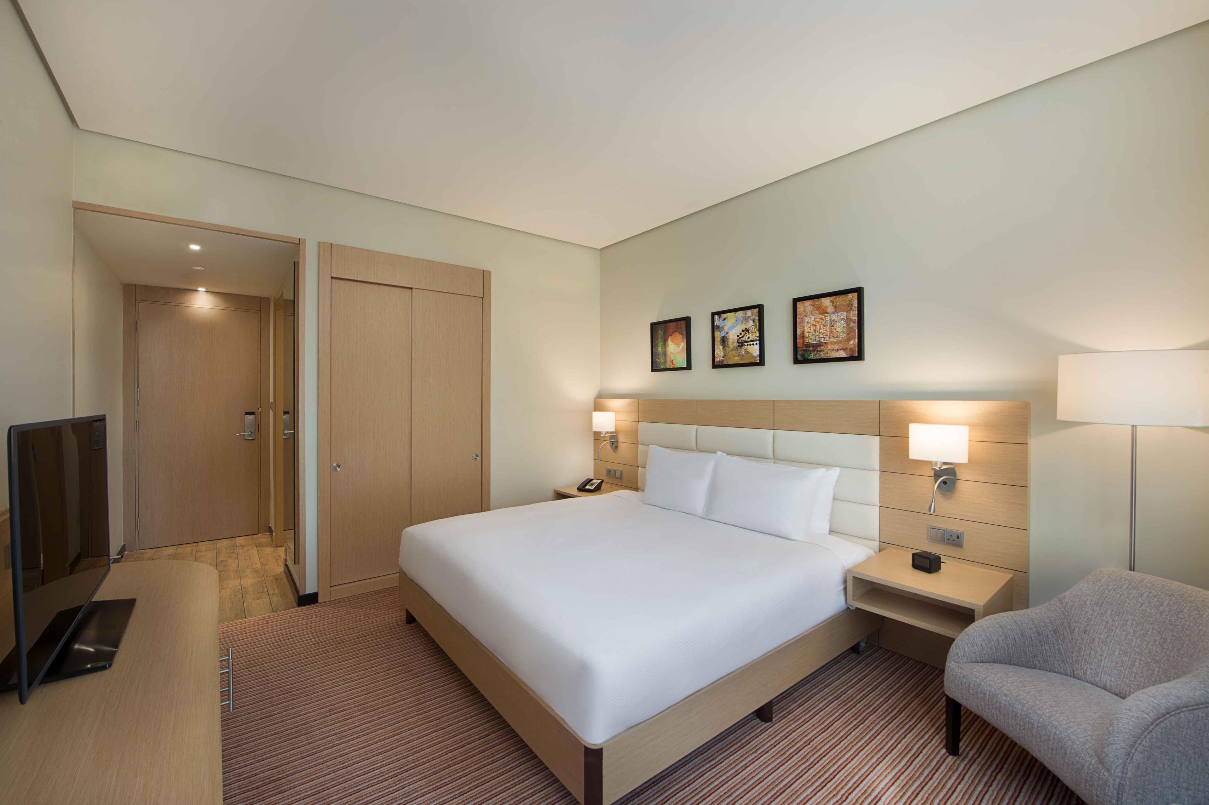 Habitación de hotel con cama, mesitas de noche, televisor, silla y una obra de arte en la pared.