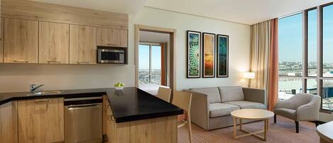 A modern hotel room with a kitchenette, a seating area with a sofa and a small table, and large windows offering a city view.