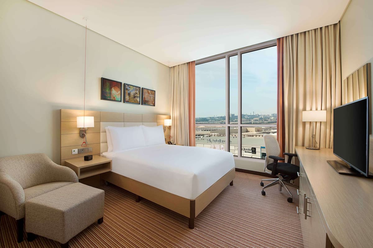 A hotel room with a large bed, a desk with a TV, a chair, a nightstand, and a view of the city through large windows.