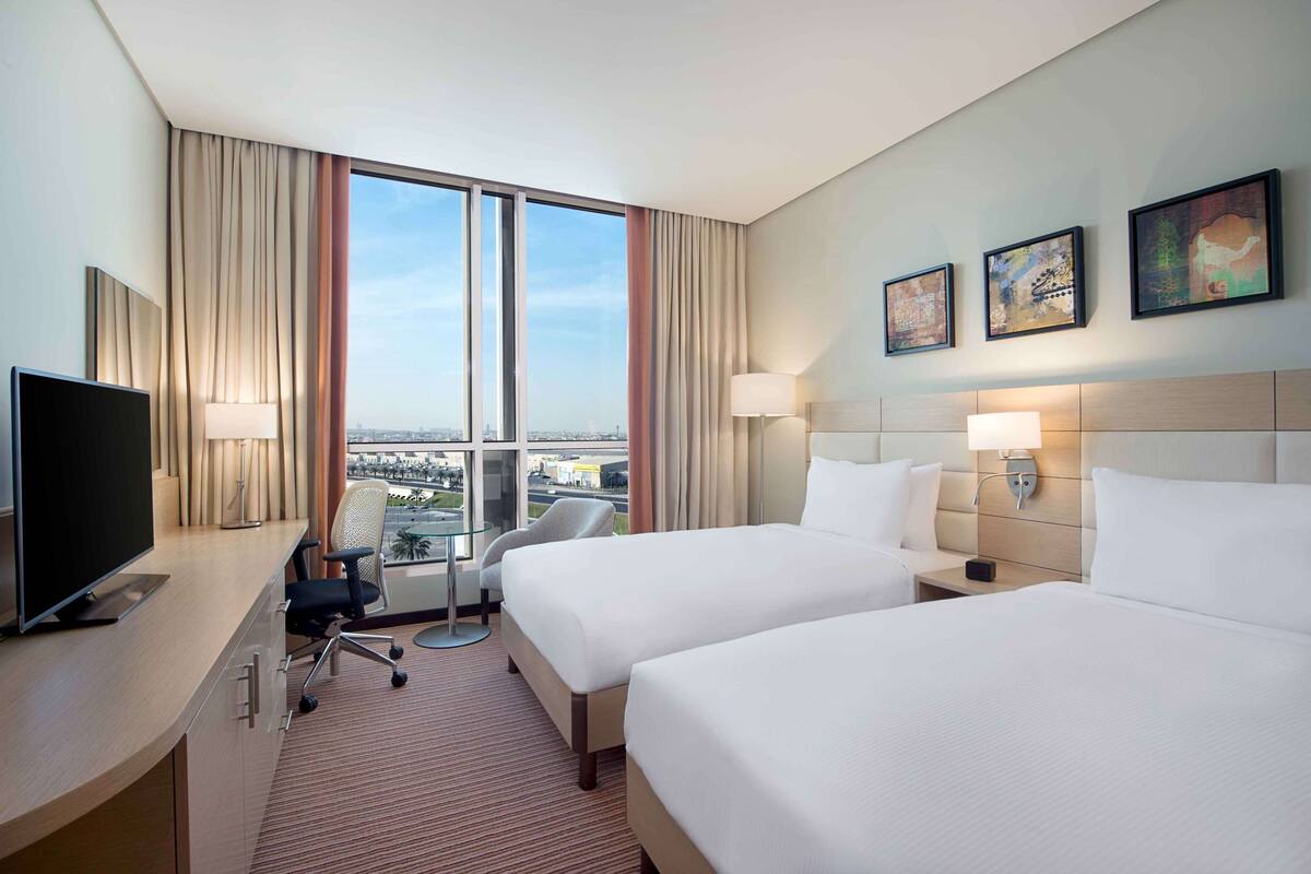 A hotel room with two beds, a desk with a TV, a chair, and a view of the city through large windows.