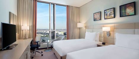 Quarto de hotel com duas camas, uma escrivaninha com televisão, uma cadeira e vista para a cidade através de janelas amplas.