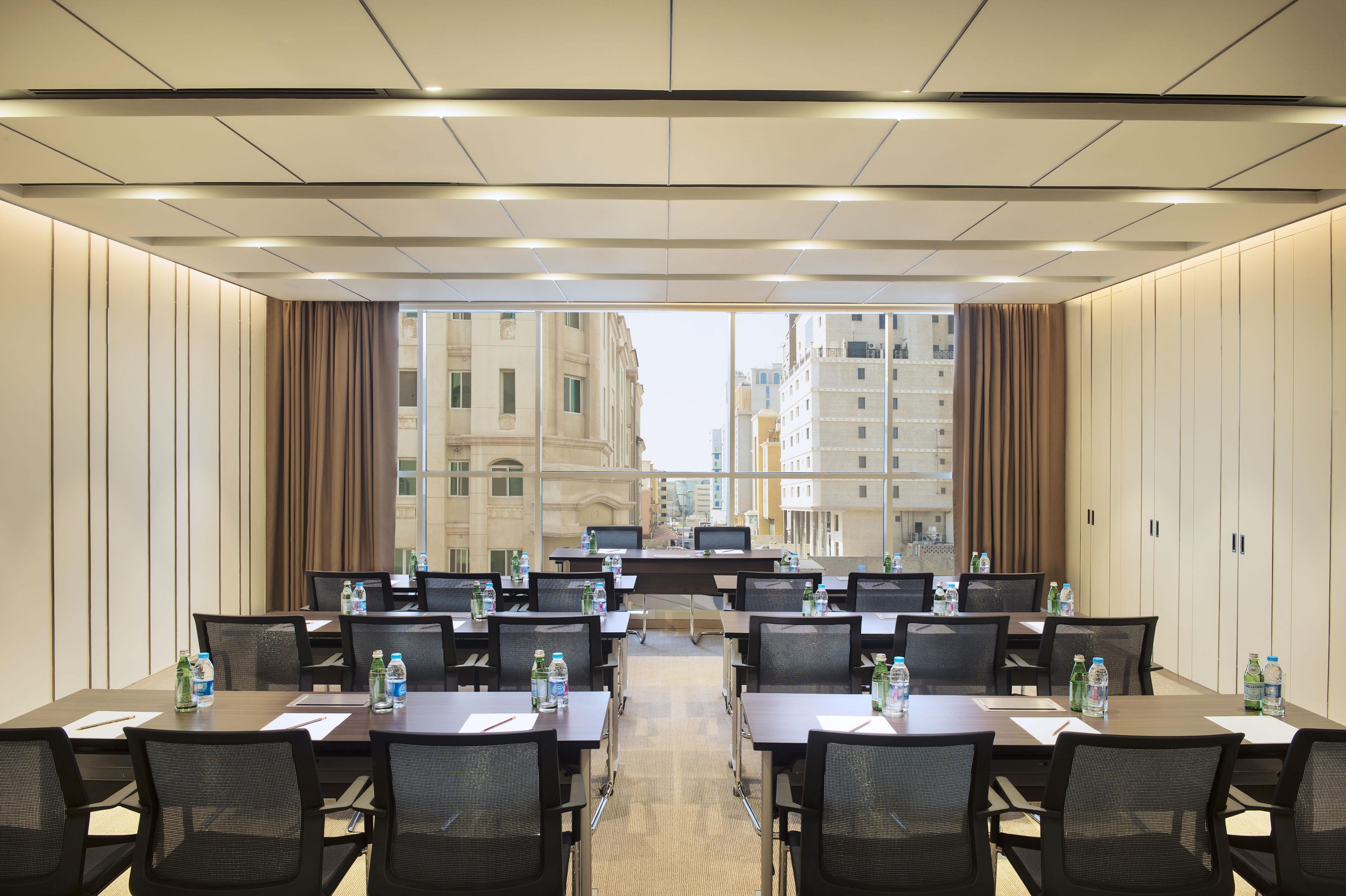 A conference room with multiple tables and chairs, equipped with water bottles and pens, and large windows offering a view of buildings.