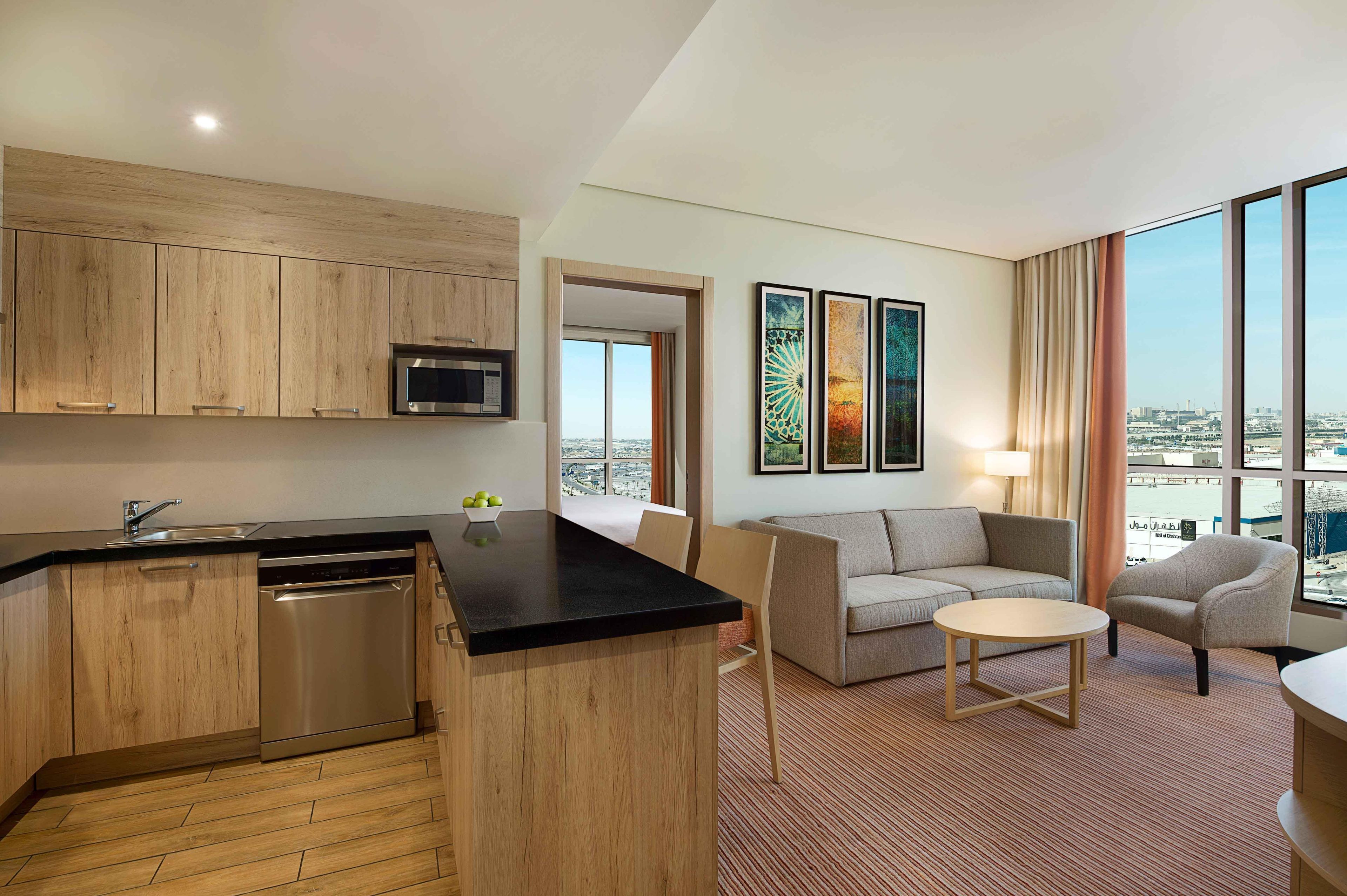 A modern hotel room with a kitchen area, a seating area with a sofa and a small round table, and large windows offering a city view.