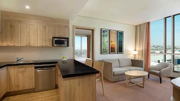 A modern hotel room with a kitchen area, a seating area with a sofa and a small round table, and large windows offering a city view.