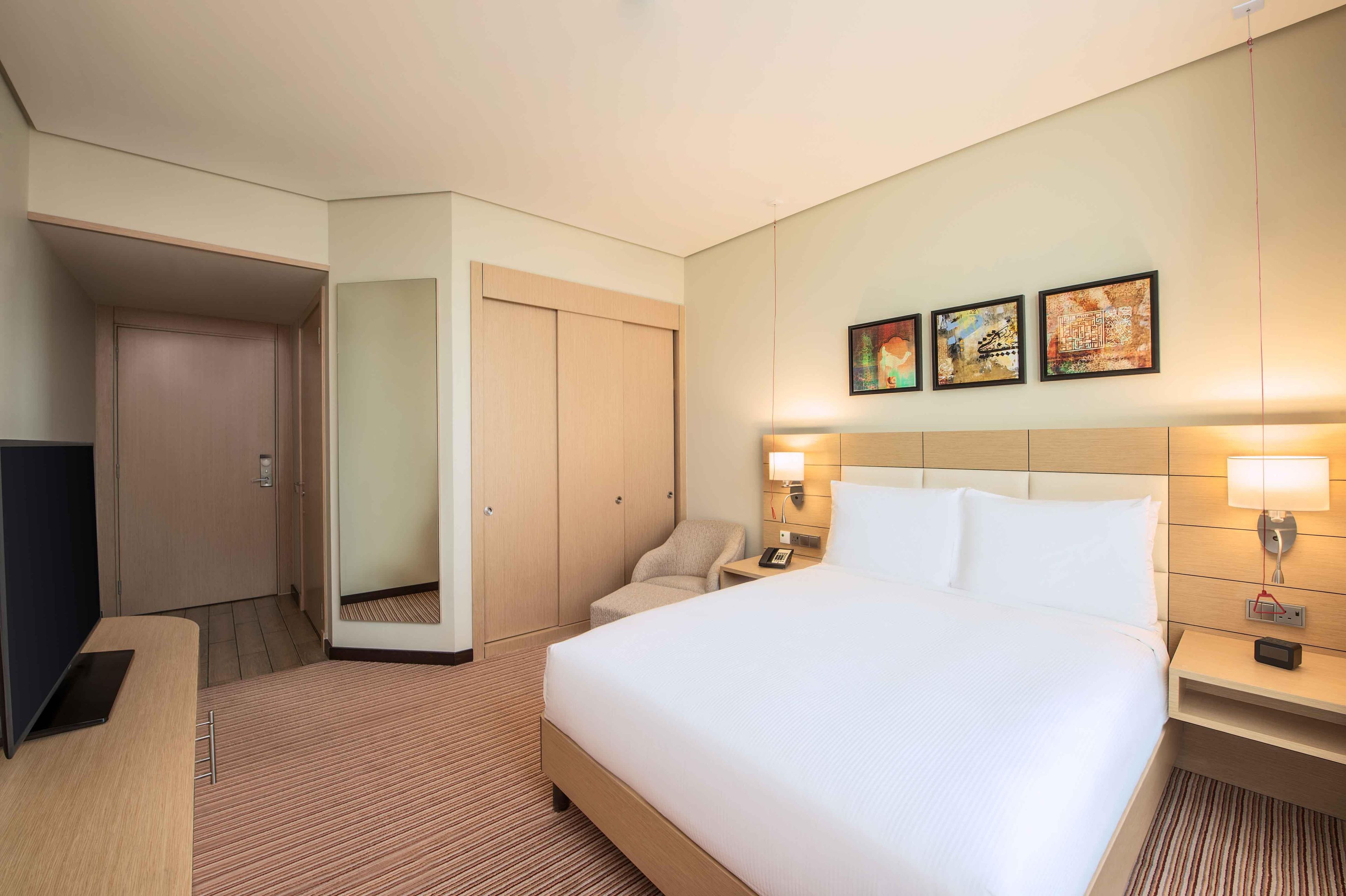 A hotel room with a bed, bedside tables, a desk, a chair, a wardrobe, and artwork on the wall.