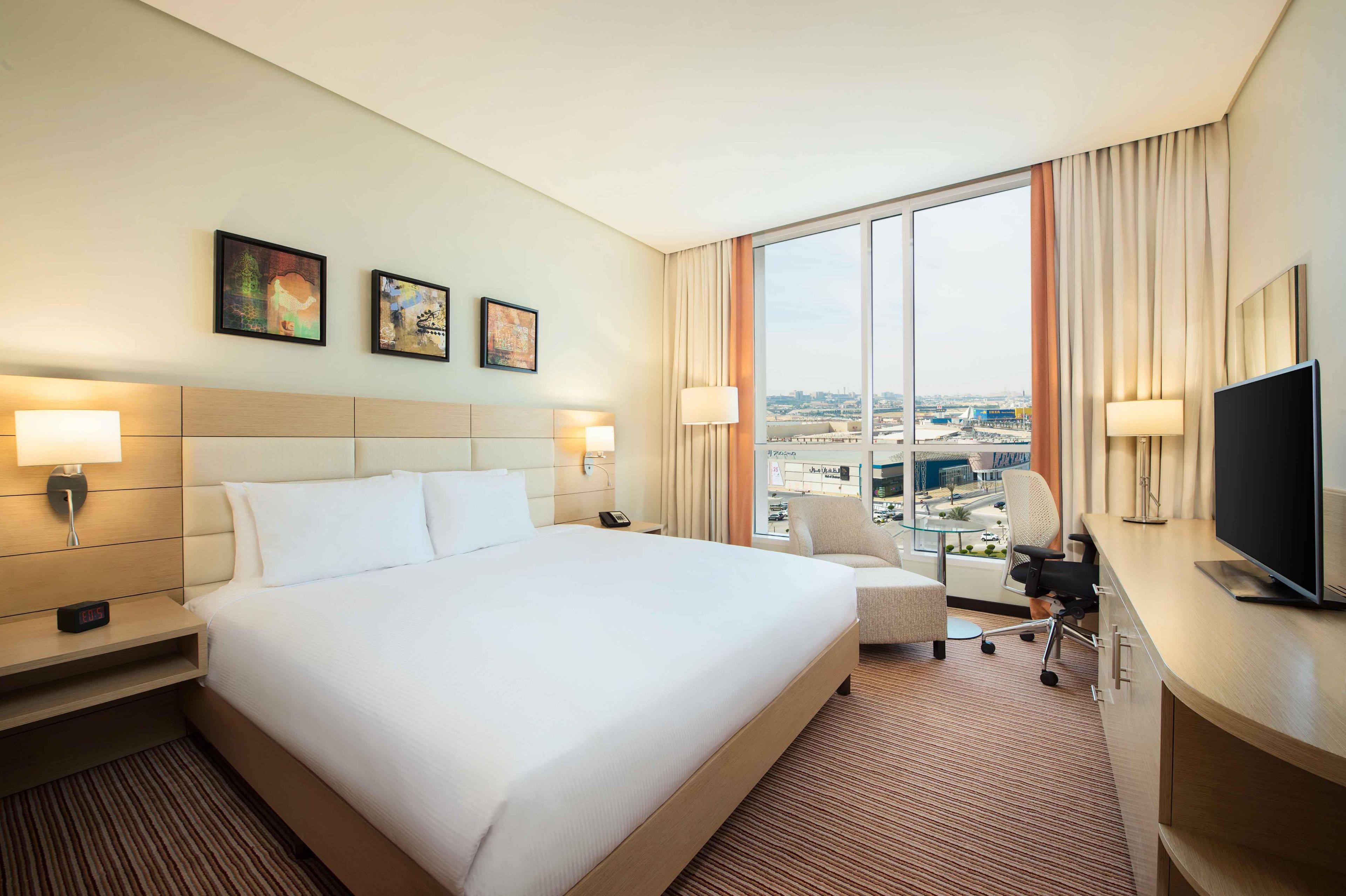 Une chambre d’hôtel avec un grand lit, deux tables de chevet avec des lampes, un bureau avec un téléviseur à écran plat, une chaise et une vue sur la ville grâce à de grandes fenêtres.