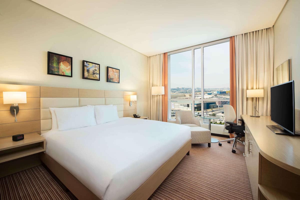 A hotel room with a large bed, a desk with a TV, a chair, and a view of the city through large windows.