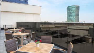 Un área de estar al aire libre con mesas y sillas, barandilla de cristal y vistas a un edificio moderno.