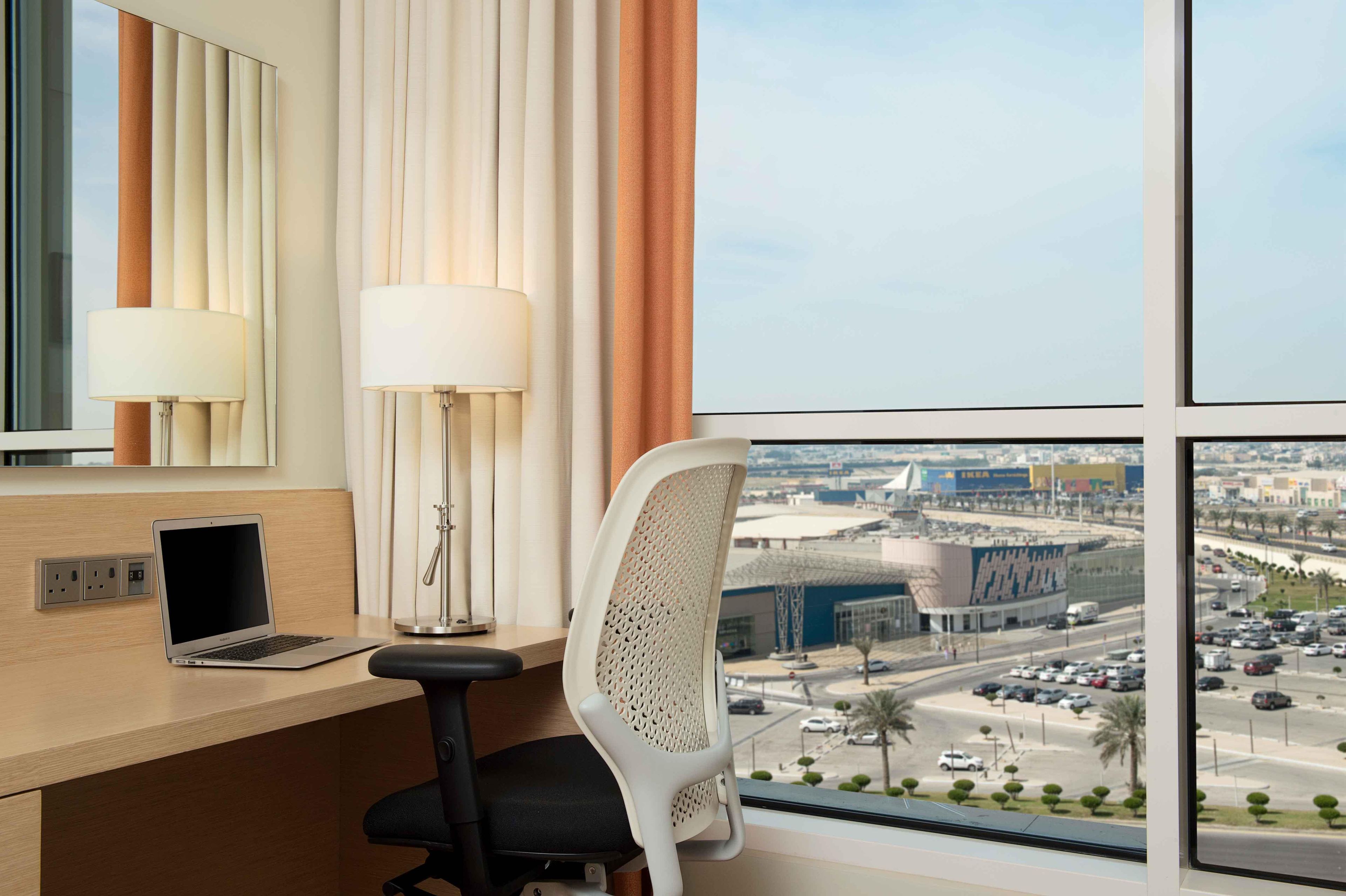 Une chambre d’hôtel équipée d’un bureau, d’un ordinateur portable, d’une lampe et offrant une vue sur le paysage urbain grâce à de grandes fenêtres.