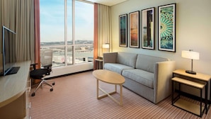 Habitación de hotel con un ventanal grande, un sofá, una mesita, un escritorio con silla y dos lámparas de pared.