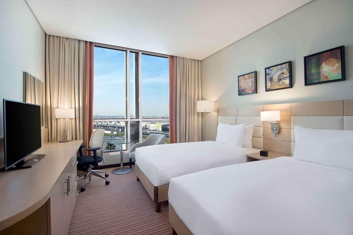 A hotel room with two beds, a desk with a TV, a chair, a lamp, and a view of the city.
