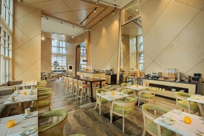 A modern dining area with multiple tables and chairs, a kitchen in the background, and large windows allowing natural light. - Hilton Garden Inn Al Khobar (Al Khobar)