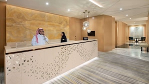Zona de recepción de hotel con pared de mármol, un mostrador y una sala de espera con dos sillones.