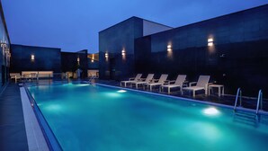 Una piscina con sillas de descanso e iluminación, rodeada de edificios en tonos oscuros.