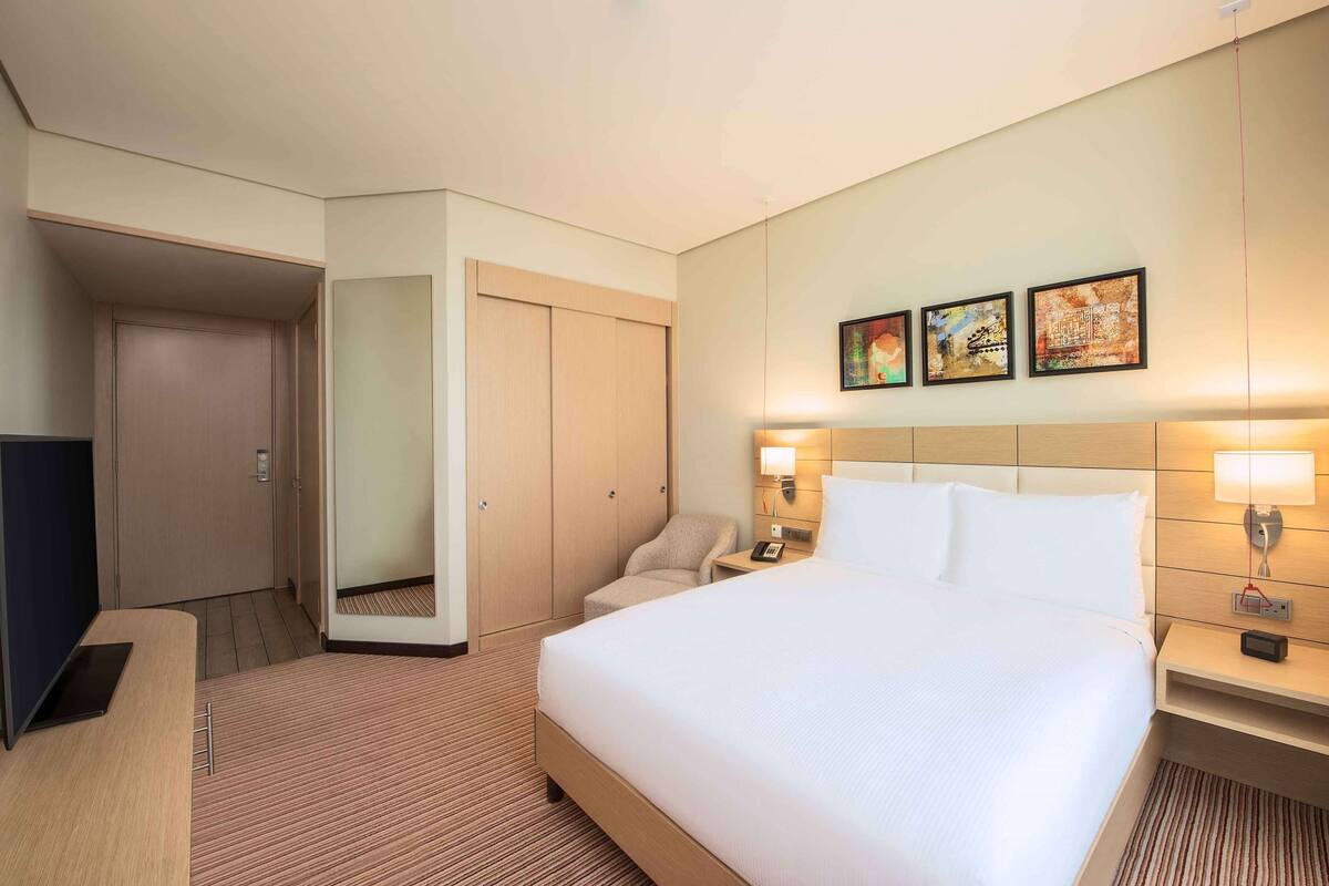 A hotel room with a large bed, bedside tables with lamps, a desk with a laptop, a chair, and a wardrobe.