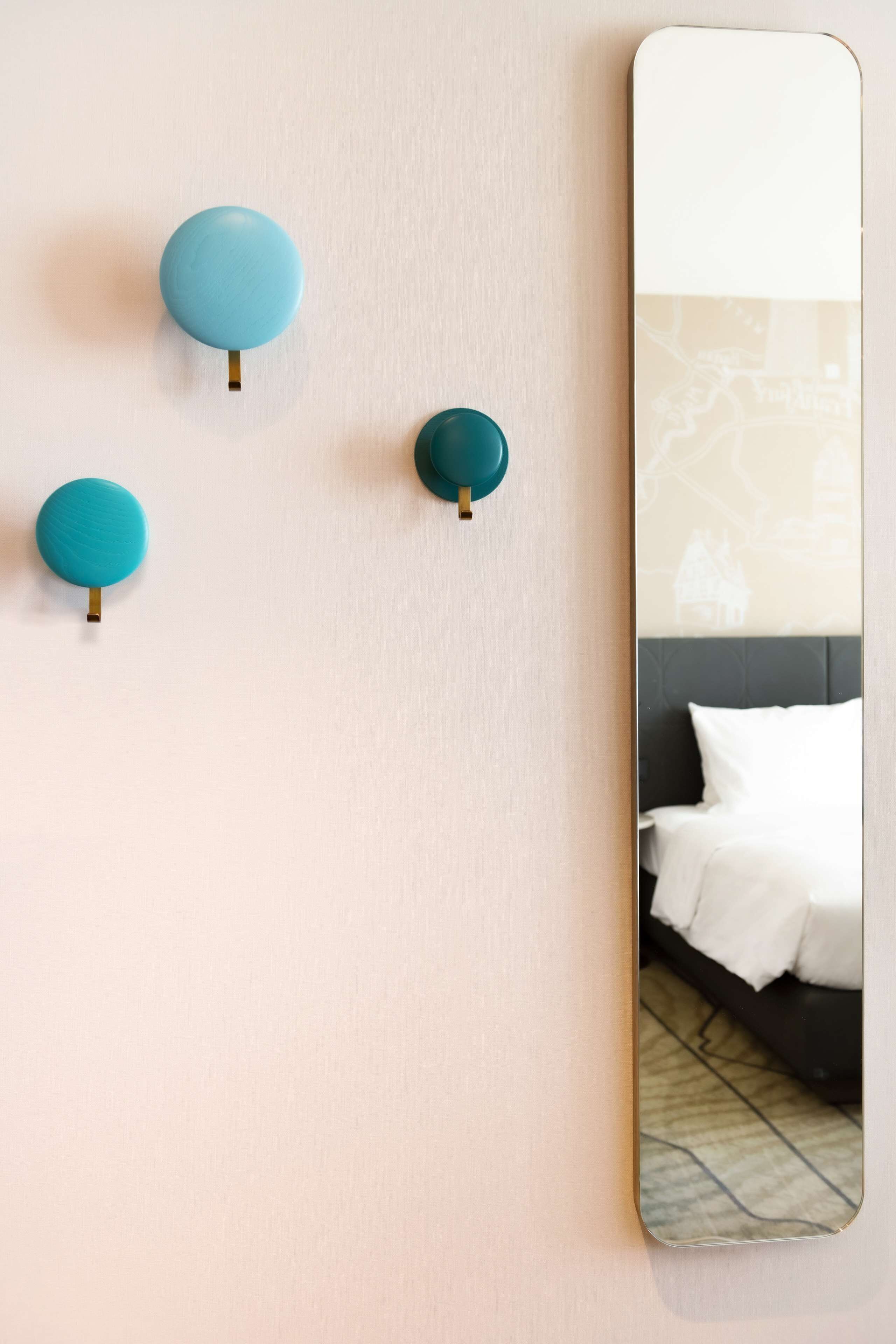 Eine Wand mit drei in Wandhalterungen montierten, türkisfarbenen Lampen und einem Spiegel, der ein Bett mit weißer Bettwäsche reflektiert.