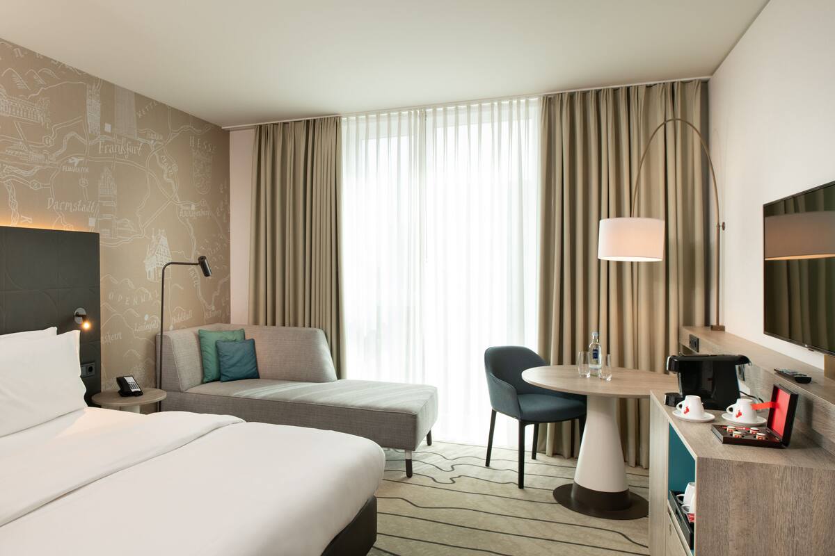 A hotel room with a bed, a sofa, a desk with a lamp, a chair, and a large window with curtains.