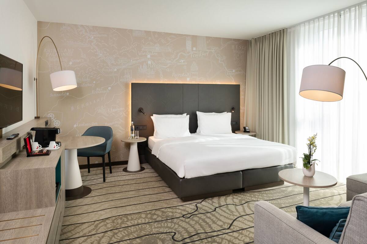 A hotel room with a large bed, a desk with a coffee maker, a chair, a small table, and a TV.
