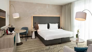 A hotel room with a large bed, a desk with a coffee maker, a chair, a small table, and a TV.