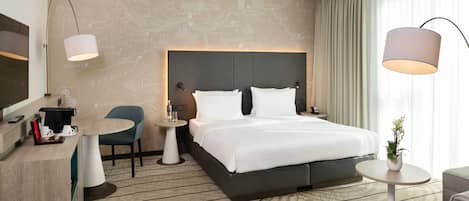 Habitación de hotel con una cama grande, un escritorio con cafetera, una silla, una mesita y un televisor.