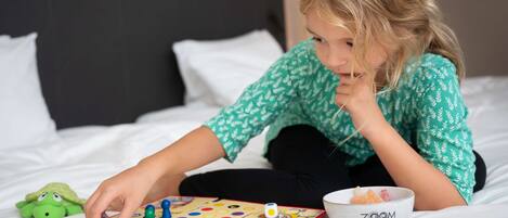 A child playing a board game on a bed with a bowl of candy and a green plush toy nearby.