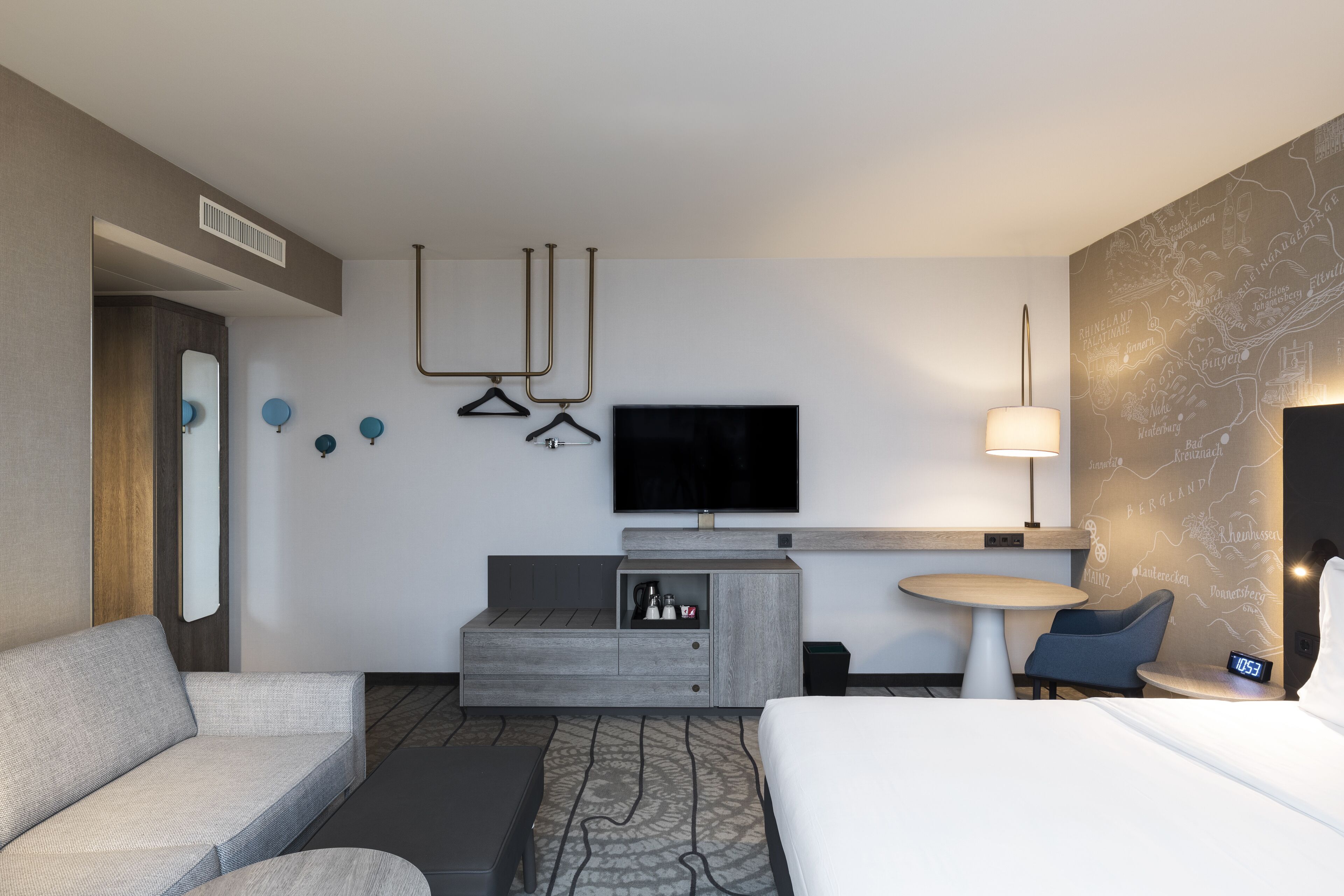 A modern hotel room with a bed, a sofa, a small table, a TV, and a wall-mounted TV.