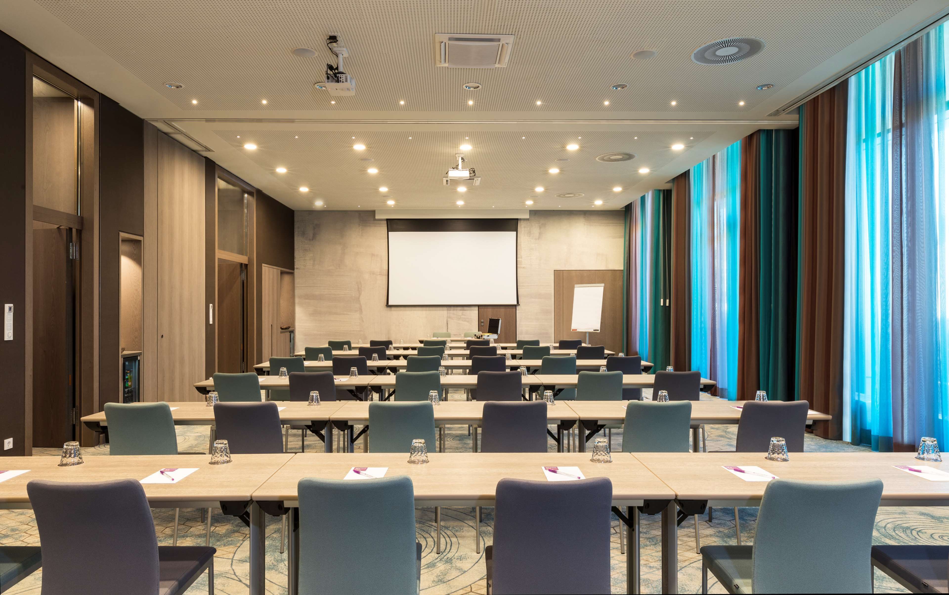 Un salón de conferencias con filas de mesas y sillas, una pantalla grande y un proyector.
