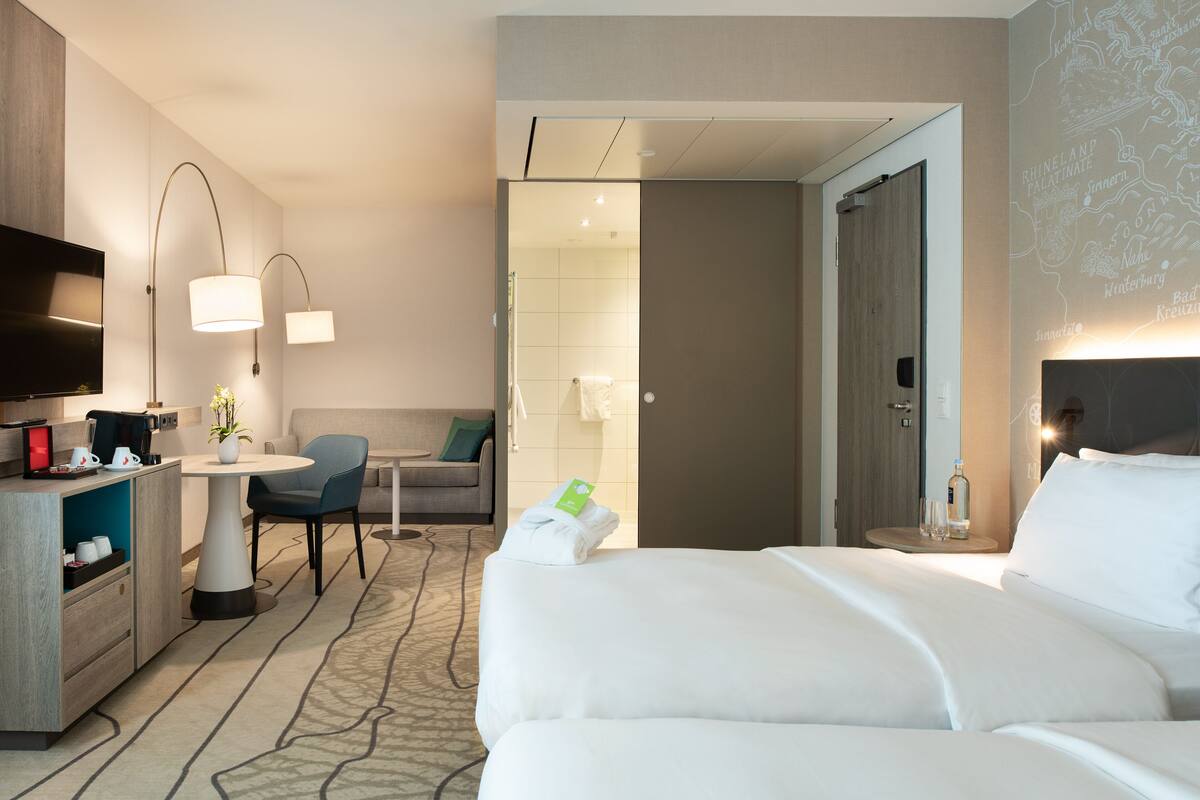 A hotel room with a bed, a desk with a chair, a television, and a wall-mounted lamp.