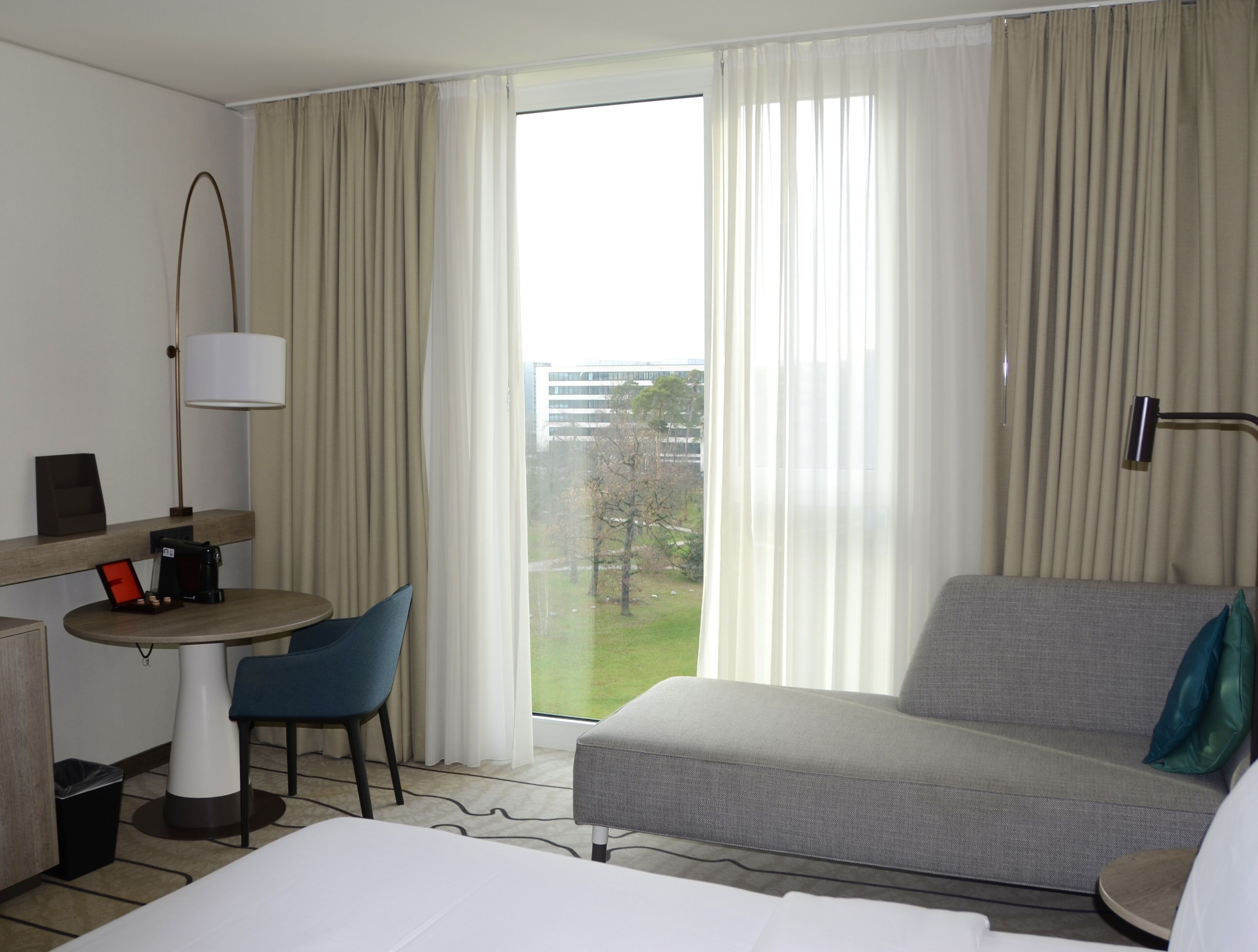 A hotel room with a bed, a sofa, a desk with a chair, a lamp, and a view of a building and trees through the window.