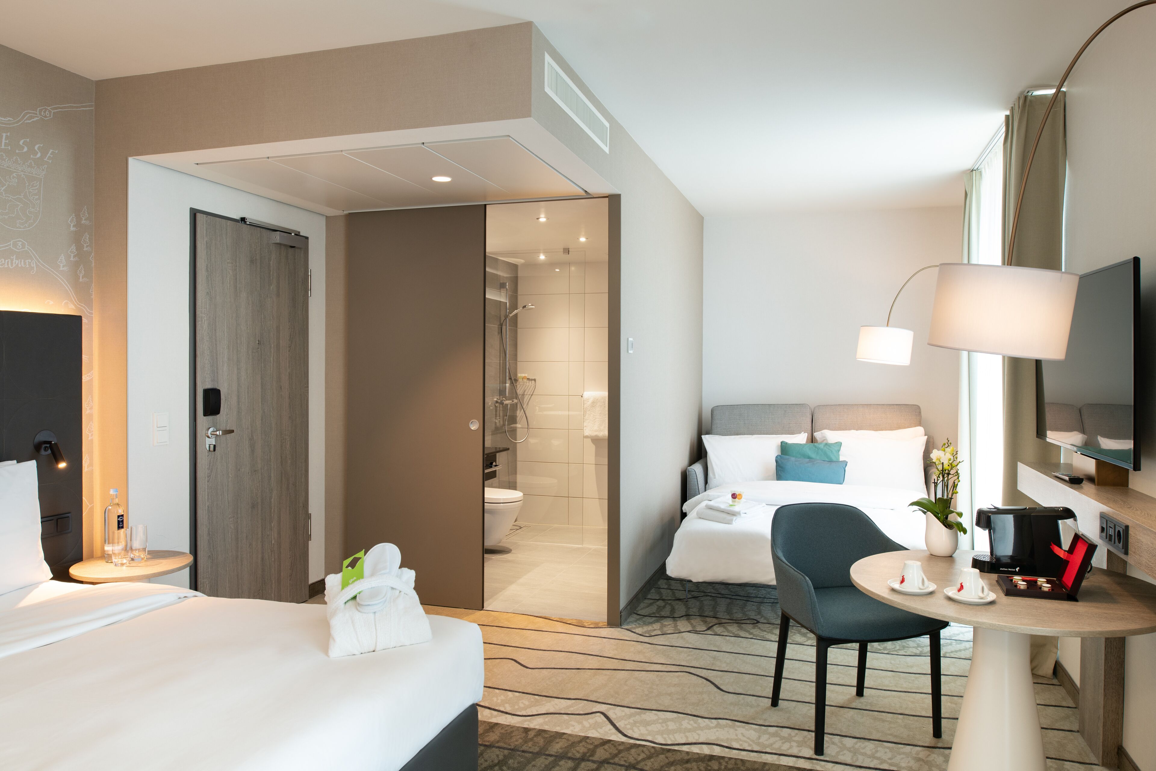 Une chambre d’hôtel avec un lit, un bureau doté d’une lampe, une chaise, un téléviseur et une salle de bain visible à travers une porte ouverte.