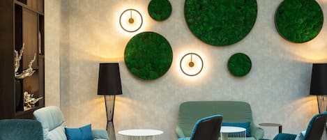 A modern lounge area with a sofa, armchairs, a round table, and a wall-mounted green decorative piece.